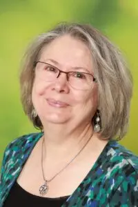 Porträt Beatrice Thiry, mit grauen Haaren, Brille und Halskette vor grünem Hintergrund