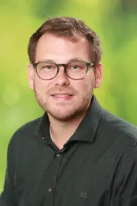 Porträt Hans-Christian Will, mit Brille und dunklem Hemd vor grünem Hintergrund