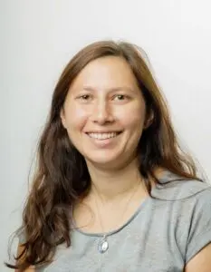 Porträt Hannelore Melcher, mit langen braunen Haaren im grauen Oberteil vor hellem Hintergrund
