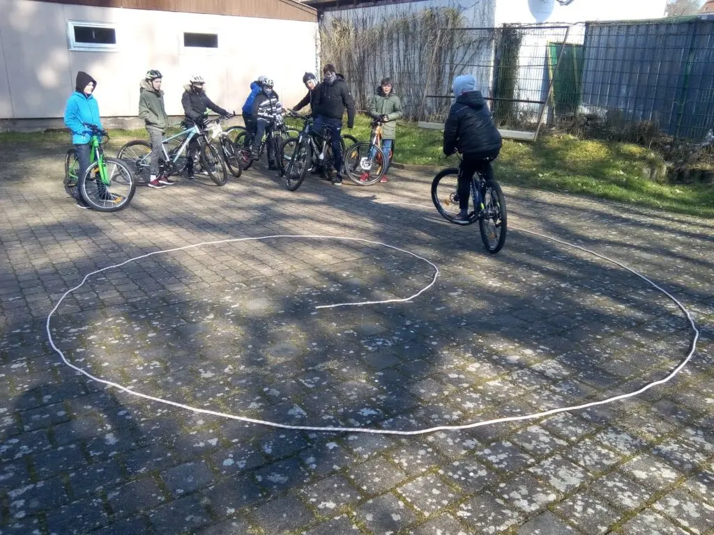 Kinder fahren mit Fahrrädern auf dem gepflasterten Schulhof im Kreis um eine aufgemalte Kreidelinie