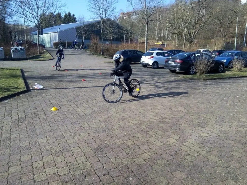 Kinder üben Radfahren auf einem gepflasterten Platz vor geparkten Autos