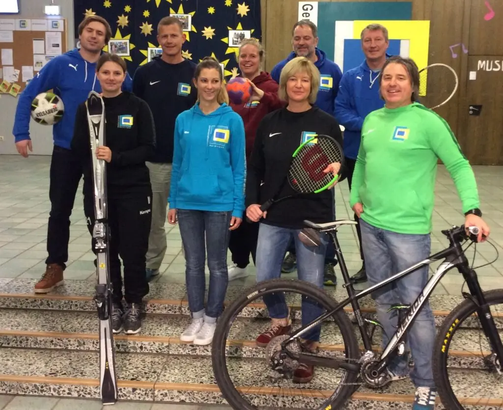 Gruppe von Jugendlichen und Erwachsenen mit Sportausrüstung, darunter ein Fahrrad und Helme, vor einer Stellwand