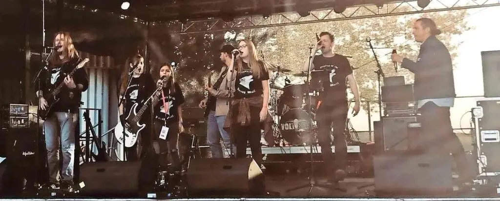Schulband spielt mit Gitarren und Mikrofonen auf einer Bühne, dahinter eine Leinwand